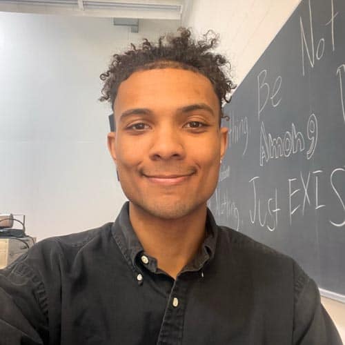 A person with curly hair and a warm smile is taking a selfie in a classroom. They are wearing a dark button-up shirt. The background shows part of a chalkboard with some text written on it, but the text is partially obscured.