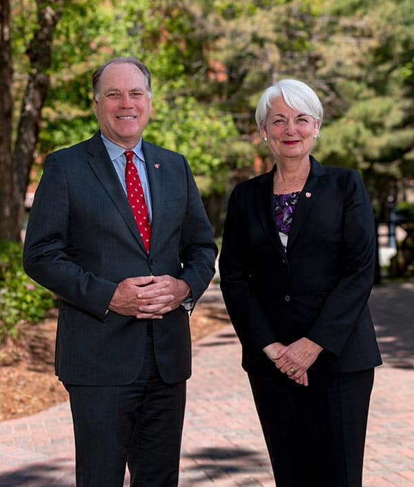 A man and a woman wearing formal business attire stand outdoors on a brick path with greenery in the background. Both are smiling at the camera. The man is in a dark suit with a red polka-dot tie, and the woman is in a dark suit with a purple patterned blouse.