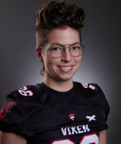 A person with short, styled hair and glasses smiles at the camera. They are wearing a black sports jersey with the number 38 and the word "Vixen" on the front. The background is dark with a subtle light halo effect behind their head.