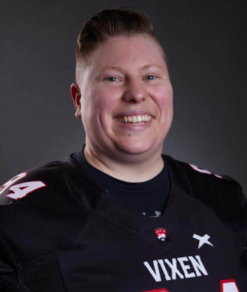A smiling person with short hair is wearing a black sports jersey with the word "Vixen" and the number "84" on it. The background is dark.