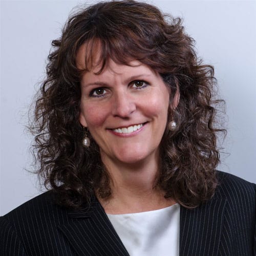 A woman with shoulder-length, curly brown hair smiles at the camera. She is wearing a dark pinstripe blazer over a light-colored blouse, and pearl drop earrings. The background is a plain light grey.