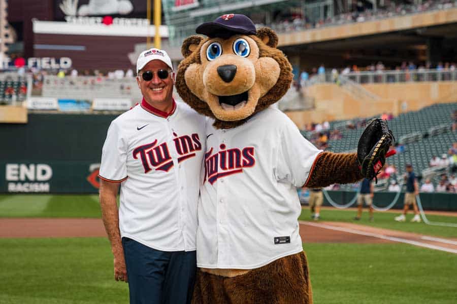 A man and a mascot bear wearing baseball jerseys and caps stand on a baseball field. The man has sunglasses and smiles with his arm around the mascot, who is holding a baseball glove. The stadium is filled with spectators in the background.