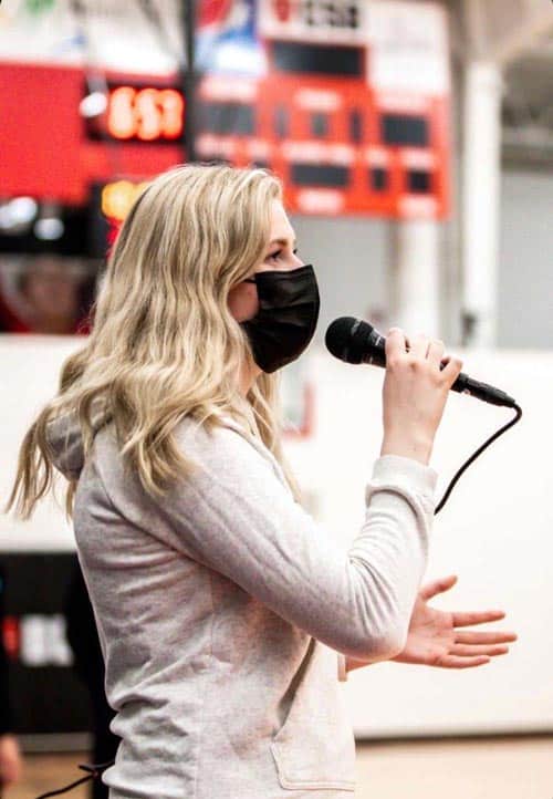A person with long blonde hair wearing a black face mask and a light-colored hoodie speaks into a microphone indoors, with an electronic scoreboard visible in the blurred background.