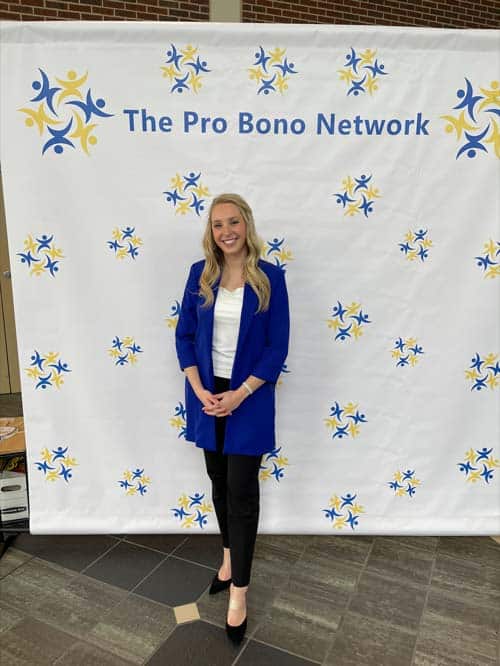A person with long hair stands smiling in front of a backdrop with "The Pro Bono Network" logo and text. They are dressed in a blue blazer, white top, black pants, and black shoes. The backdrop is decorated with yellow and blue abstract figures.