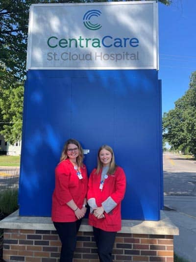 Two people smile and stand in front of a blue CentraCare St. Cloud Hospital sign. Both are wearing red jackets over black pants and have name badges clipped to their jackets. A tree and a road are visible in the background on a sunny day.