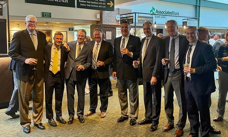 Seven men in suits stand together in an indoor setting, smiling and holding drinks. They appear to be at a social event and are posing for the photo. The background includes signs for various club sections and an Associated Bank sign on a wall.