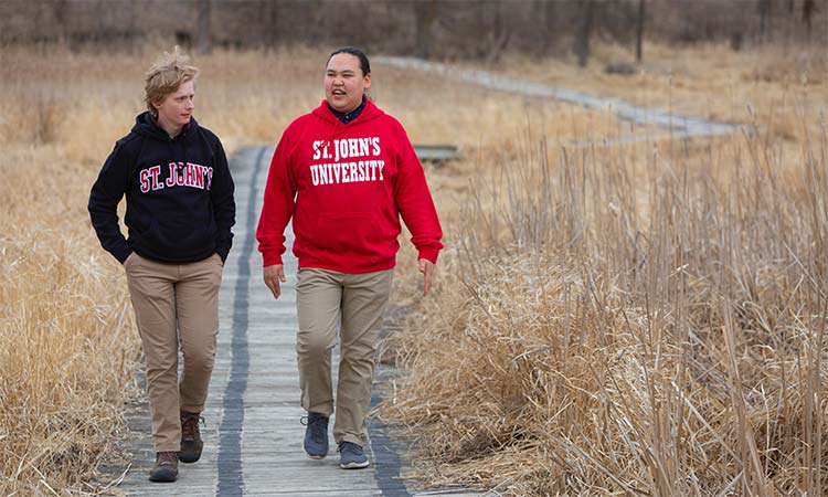Two people in St. John’s University sweatshirts walk together on a wooden boardwalk through a grassy area, engaged in conversation. One wears a red sweatshirt and the other a black one; both wear beige pants. The surrounding landscape is dry with tall grasses.