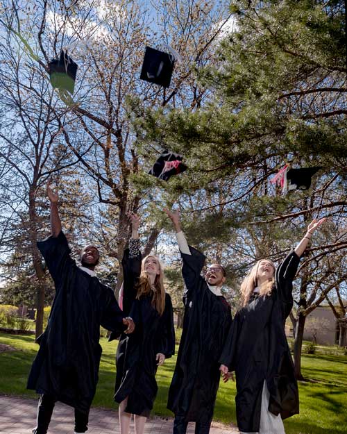 Four graduates in black robes joyfully throw their caps into the air. They're standing outdoors on a sunny day with green trees and a park-like setting in the background.