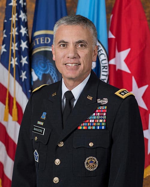 A man in a formal military uniform stands in front of several flags, including the American flag. He has short graying hair and is smiling. His uniform is adorned with various badges, ribbons, and insignia.