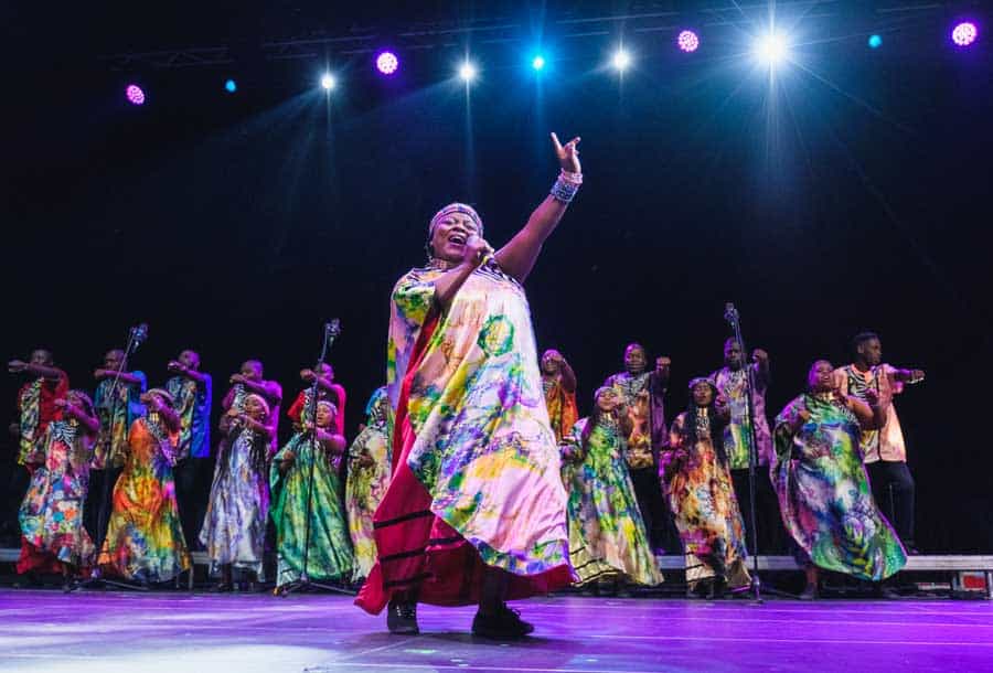 A group of performers in vibrant, colorful attire sing and dance on stage under bright stage lights. The lead performer, front and center, raises one arm and gestures energetically, creating a lively and dynamic atmosphere.