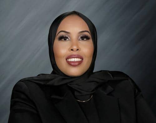 A person wearing a black hijab and black clothing smiles against a dark, gradient background. Their makeup is beautifully applied, featuring bold eye makeup and a vibrant lipstick.