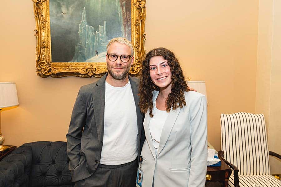 Two people stand close together, both smiling at the camera. The person on the left has short blonde hair, glasses, and a beard, wearing a light gray suit and white t-shirt. The person on the right has long curly hair, wearing a light gray suit and white shirt. They are in a room with an ornate framed painting and lamps in the background.