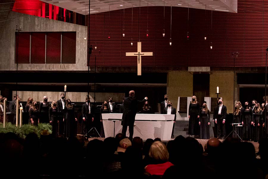 A choir dressed in black performs in a church with a large cross at the center. A conductor stands in front, leading the singers. The audience is seated, watching the performance. The church's interior is dimly lit, with accents of red and candles on the altar.