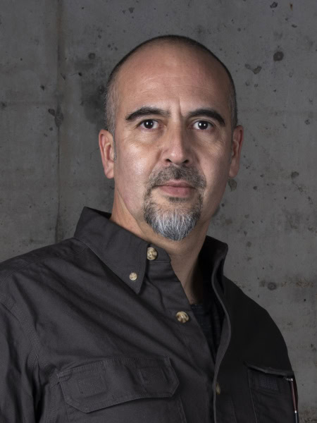 A middle-aged man with a bald head and a gray goatee is wearing a dark button-down shirt. He is standing against a gray, textured concrete wall. His expression is neutral as he looks directly at the camera.
