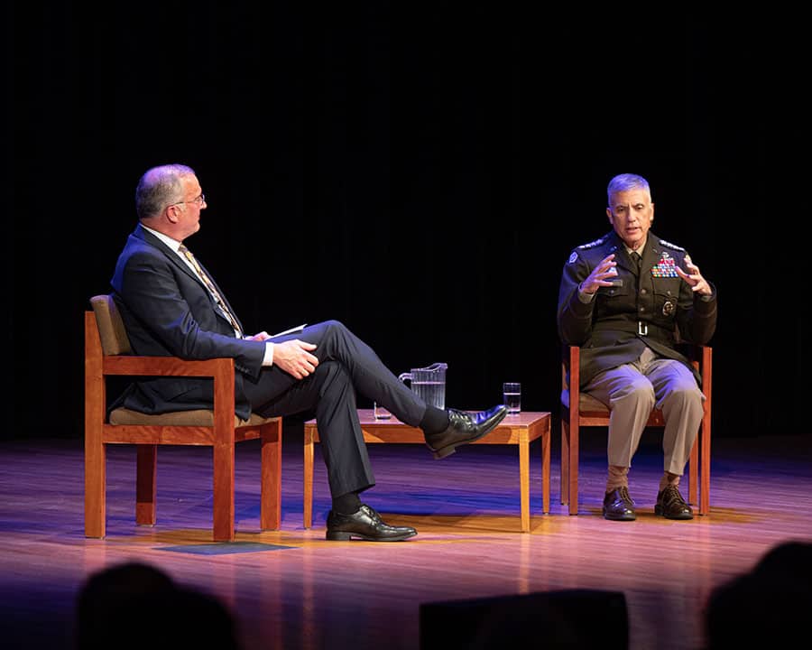 Two men are seated on stage in wooden chairs during a discussion. The man on the left is in a suit and glasses, while the man on the right is in a military uniform. Both are engaged in conversation, with the man in uniform gesturing with his hands. A small table is between them.