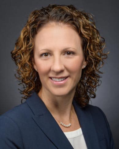 A woman with curly brown hair and fair skin smiles at the camera. She is wearing a dark blazer, a white blouse, and a pearl necklace. The background is plain and dark.