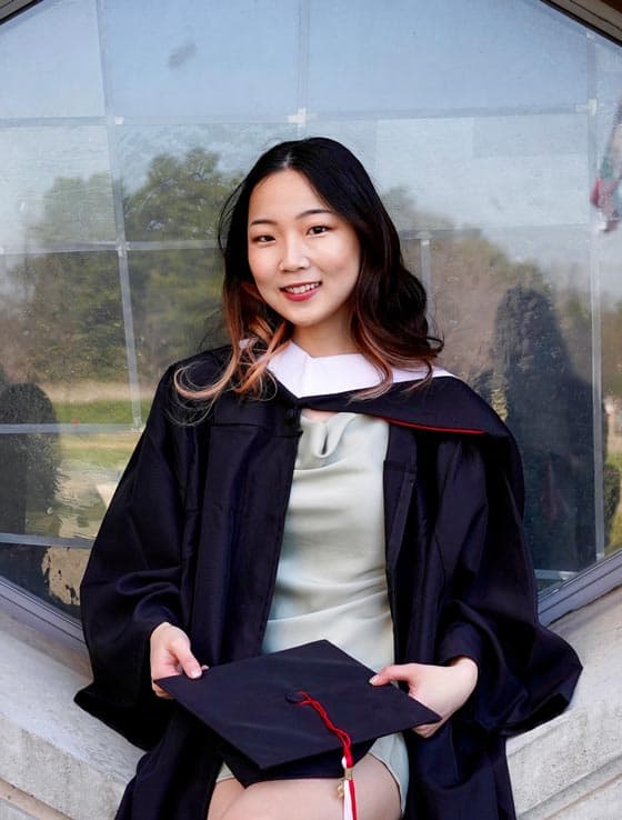 A young woman is sitting in front of a window wearing a graduation cap and gown. She is holding her cap in her lap and smiling at the camera. The background is blurred, showing some greenery and parts of the building's exterior.