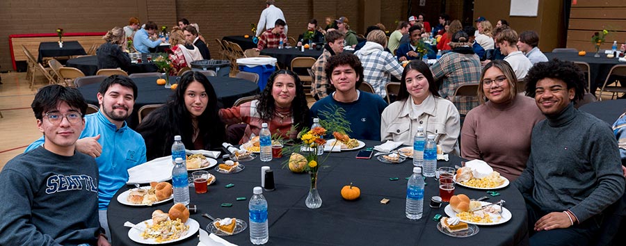 A diverse group of eight young adults sitting around a table in a large dining hall, sharing a meal. The table is adorned with water bottles, plates of food, and small decorative pumpkins. Other groups of people are seen dining in the background.