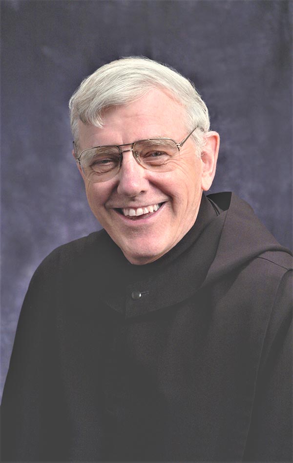 A smiling older man with short gray hair and glasses, dressed in a dark robe, poses against a dark backdrop.