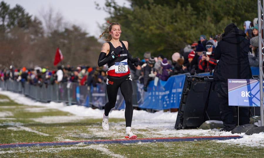 A woman wearing athletic gear and a bib number 605 is running across the finish line at an outdoor race event. There is snow on the ground and a crowd of spectators behind barriers, some taking photos or cheering. A sign indicates the race distance as 8K.