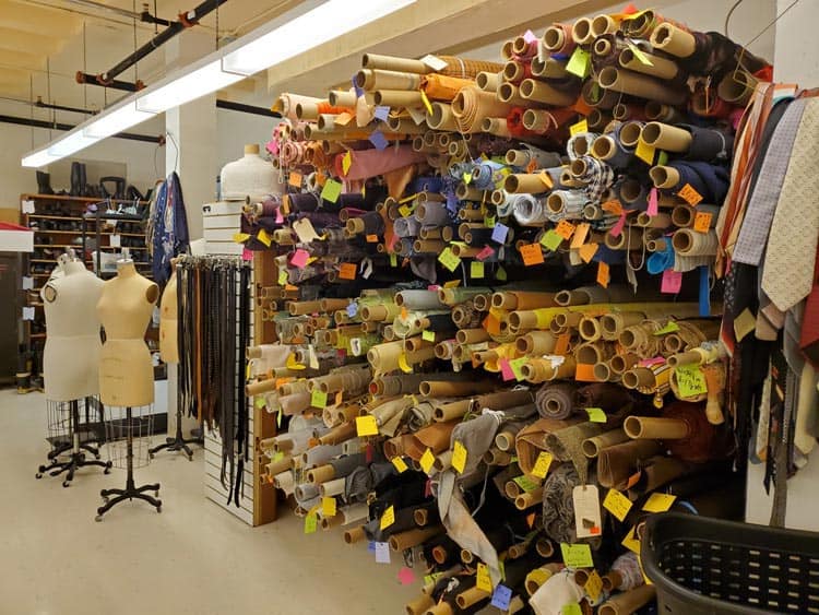 A fabric store displays numerous rolls of fabric on wall shelves, each labeled with colorful tags. Mannequins stand nearby, and various sewing materials are visible in the background, creating a bustling and creative atmosphere.