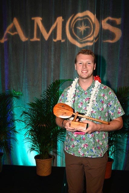 A person wearing a green floral shirt and a white lei stands smiling while holding a wooden award. The background features potted plants and a backdrop with the text "AMOS" projected on it.