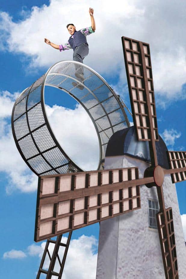 A person is balancing on top of a windmill that has four wooden blades and a large circular metallic turning mechanism. The sky is bright blue with scattered clouds in the background. The individual seems to be performing a stunt as they stand confidently on the windmill.