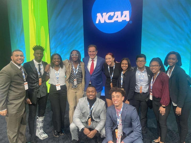A group of people in business attire stand in front of an NCAA backdrop. They are smiling and appear to be at a formal event or conference. The group consists of two rows, with some standing and others kneeling or crouching in the front.