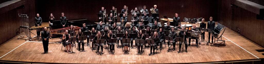A symphonic band posed on a wooden stage in a concert hall. Members are dressed formally in black attire, each with their respective instruments, including wind, brass, and percussion sections, arranged in tiers. The conductor stands at the left front.
