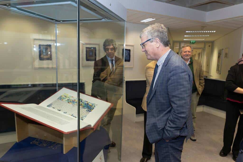 A man in a suit examines a large open book displayed in a glass case at an exhibition. Two other people are seen in the reflection of the glass, observing the exhibit as well. The setting appears to be a modern, well-lit room, possibly a museum or gallery.