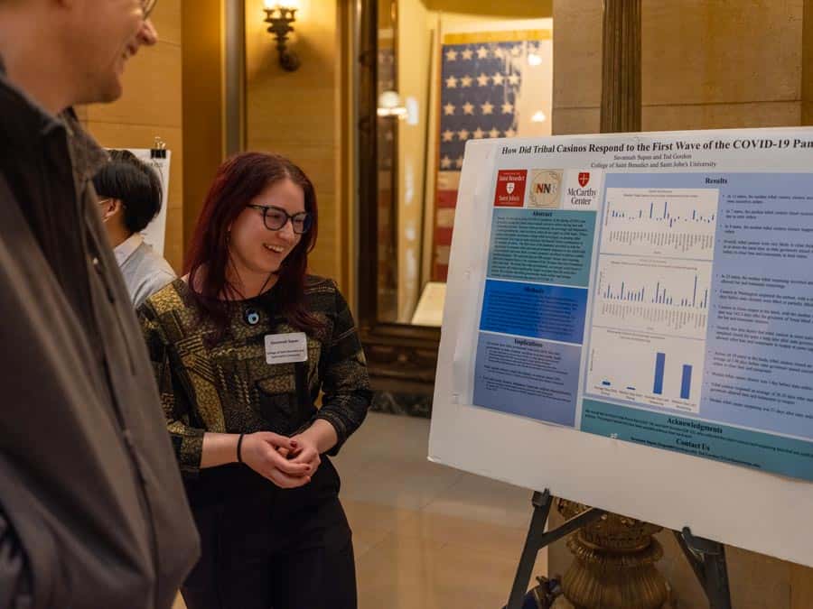 A woman with glasses and long dark hair smiles while presenting a research poster titled "How Did Tribal Casinos Respond to the First Wave of the COVID-19 Pandemic?" to a man in a suit. The poster features text, graphs, and charts. An American flag is in the background.