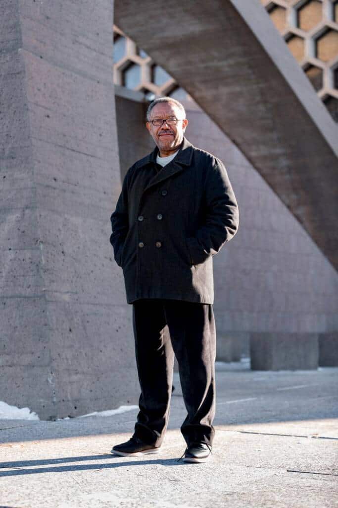 A man stands outdoors in front of a large concrete structure with a honeycomb pattern. He is wearing a black coat, black pants, and glasses, with his hands in his coat pockets. The sun is shining, casting shadows.