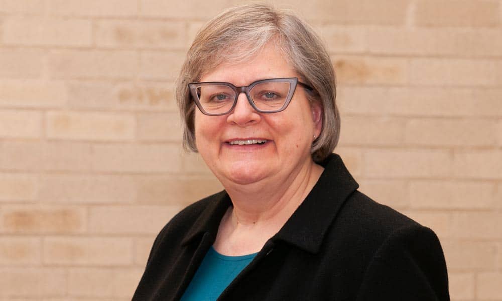 A smiling person with short gray hair and glasses stands in front of a beige brick wall. They are wearing a dark blazer over a teal top.