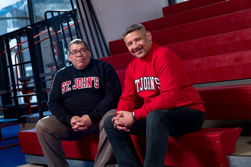 Two men are sitting side by side on red bleachers. Both are wearing St. John's sweatshirts—one in black and the other in red. The background features a staircase and a window revealing an outdoor view. They are smiling and appear relaxed.