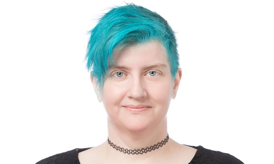 A person with bright turquoise short hair is looking straight at the camera, smiling subtly. They are wearing a black choker necklace and a dark top. The background is plain white.