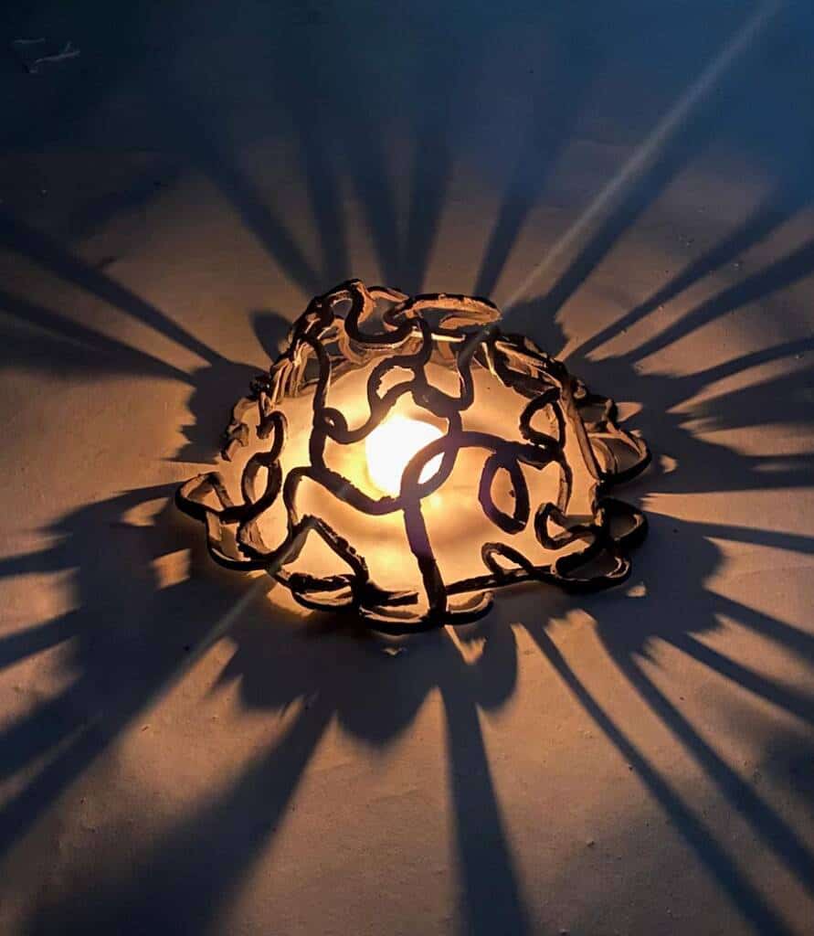 A glowing light bulb encased in a decorative, abstract metal frame casts intricate shadows on the surrounding surface. The shadow patterns radiate outward, creating a starburst effect on the ground.