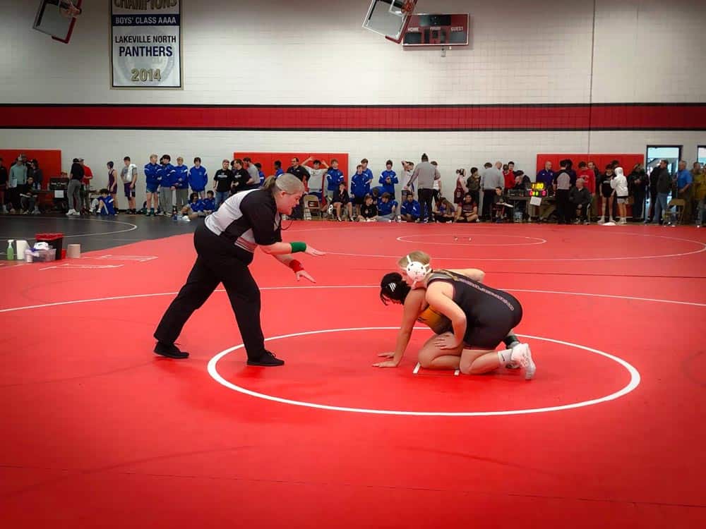 A wrestling match unfolds in a gym with red mats. One wrestler, dressed in black, is on their hands and knees while the referee kneels, giving instructions. Spectators, including a group in blue uniforms, watch from the edges of the gymnasium.
