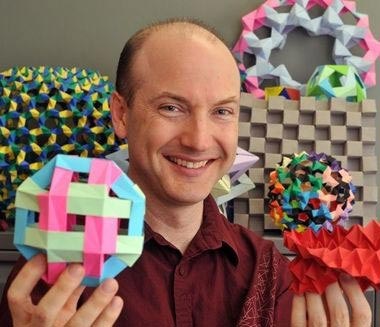 A person with a big smile holds two colorful origami geometric shapes in their hands. Behind them, various intricate and brightly colored origami structures are displayed on a showcase. They are wearing a maroon shirt.