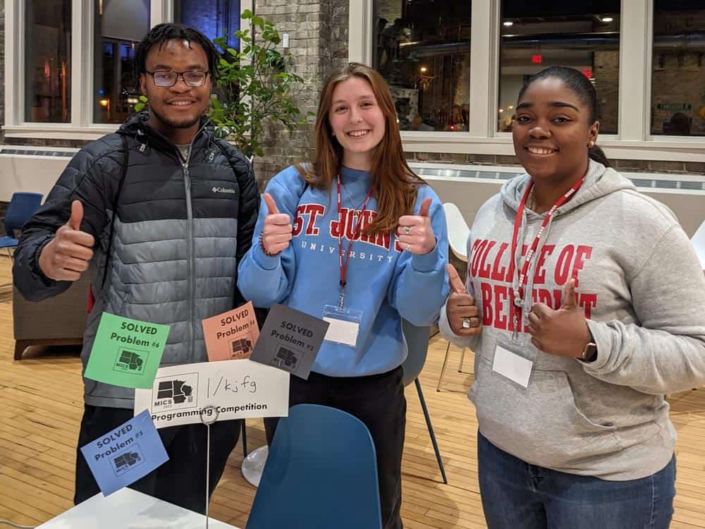 Three individuals smile and give thumbs up at a programming competition. They stand behind a table with a sign and colorful paper tags reading "SOLVED Problem #1" and "#2." One person wears a puffer jacket, another a "St. John's" sweater, and the last a "College of DuPage" sweatshirt.