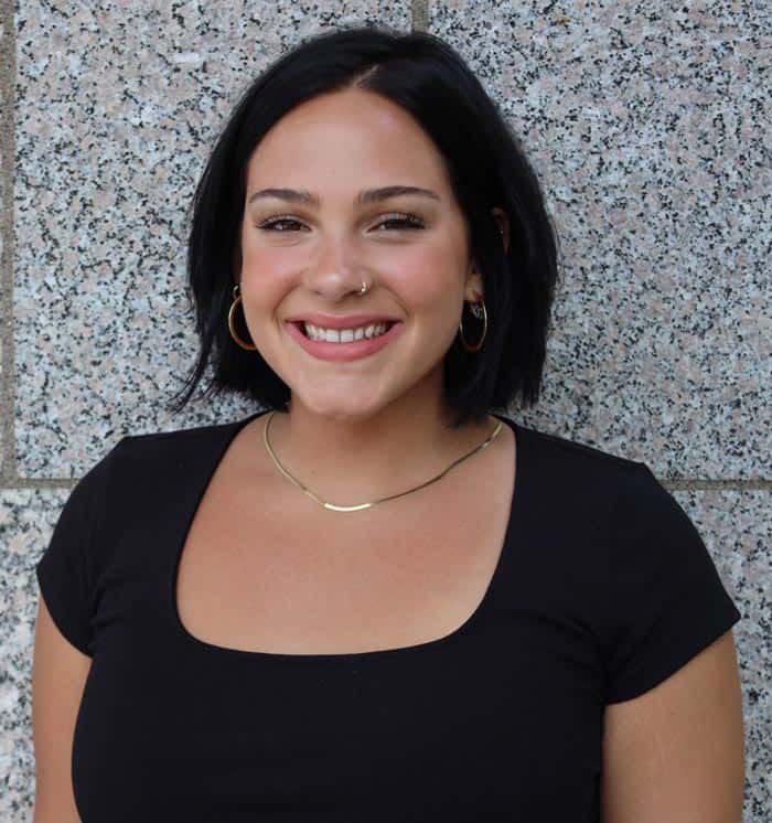 A woman with shoulder-length black hair is smiling at the camera. She is wearing a black top, gold hoop earrings, a delicate gold necklace, and has a nose piercing. The background is a speckled gray textured wall.