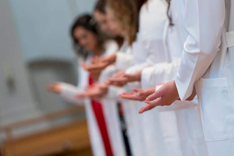 A group of individuals wearing white lab coats stands in a line with hands extended forward in a gesture. The background is blurred, focusing on the hands and arms of the people.