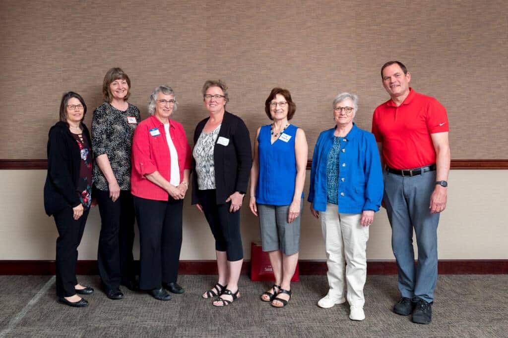 A group of seven people stands indoors in a line facing the camera. Four women are wearing name tags, and the individuals are dressed in casual attire. The background is a plain wall with a textured surface and a carpeted floor.