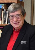 A smiling person with short gray hair and glasses, dressed in a black blazer with a red shirt underneath. They are standing in front of a bookshelf filled with books.