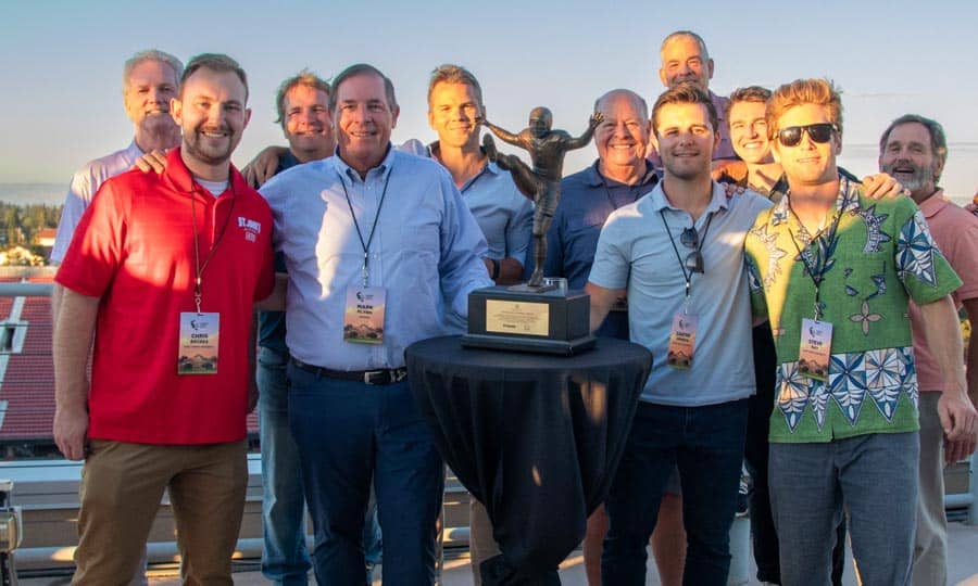 A group of people stands outdoors around a tall table with a trophy, smiling at the camera. They wear lanyards and casual attire, with a clear sky in the background. The atmosphere appears celebratory.