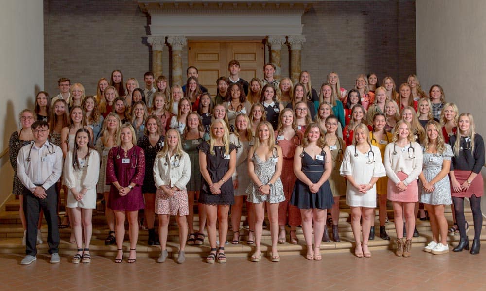 A large group of medical students is gathered indoors, standing on stairs and posing for a group photo. Many of them are wearing stethoscopes, name tags, and professional attire, suggesting a formal event or ceremony. The background features a brick wall and wooden door.