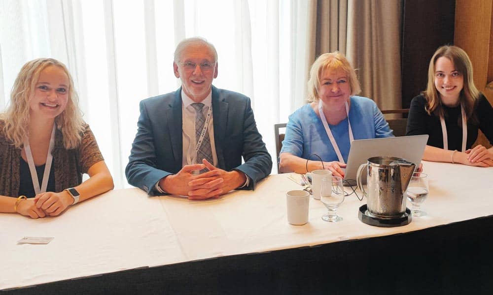 Four people are sitting at a conference table with a white tablecloth. From left to right, there are two women, a man in a suit, and another woman with a laptop. They are all smiling at the camera.