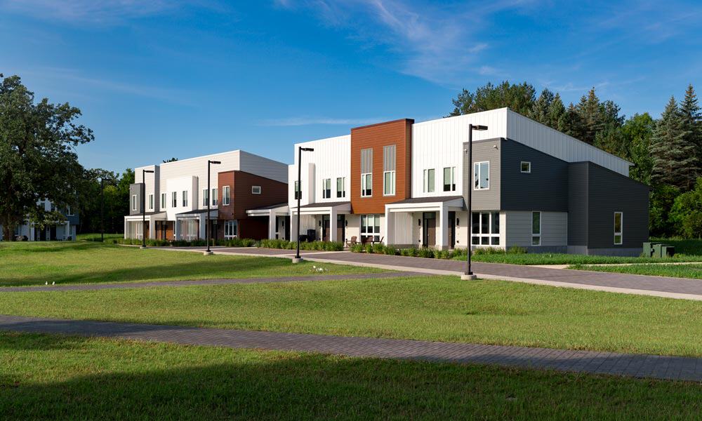 A row of modern, two-story townhouses with clean lines and large windows. The houses feature white and gray exteriors with wooden accents. A manicured lawn and paved walkway extend in front of the homes, with trees and a blue sky in the background.