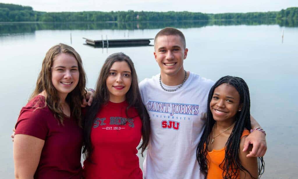 Four young adults stand closely together and smile near a calm lake with a floating dock in the background. They are wearing casual clothes, including t-shirts with college logos, and the scene is bright and peaceful with trees lining the horizon.