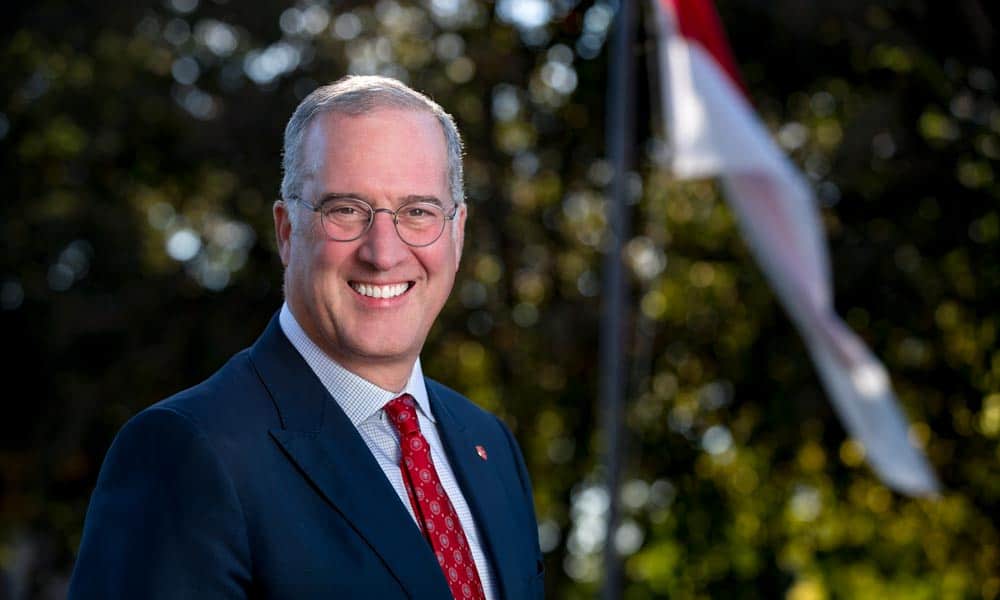 A man wearing glasses, a suit, and a red tie smiles at the camera. He stands outdoors with a blurred background of trees and a partially visible flag. The lighting highlights his face and the top of his head.