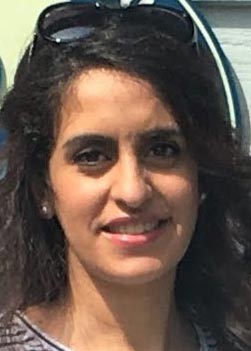 A person with long dark hair is smiling at the camera. They are wearing sunglasses on top of their head and a pair of earrings. The background is partially visible, showing some blue and white objects.